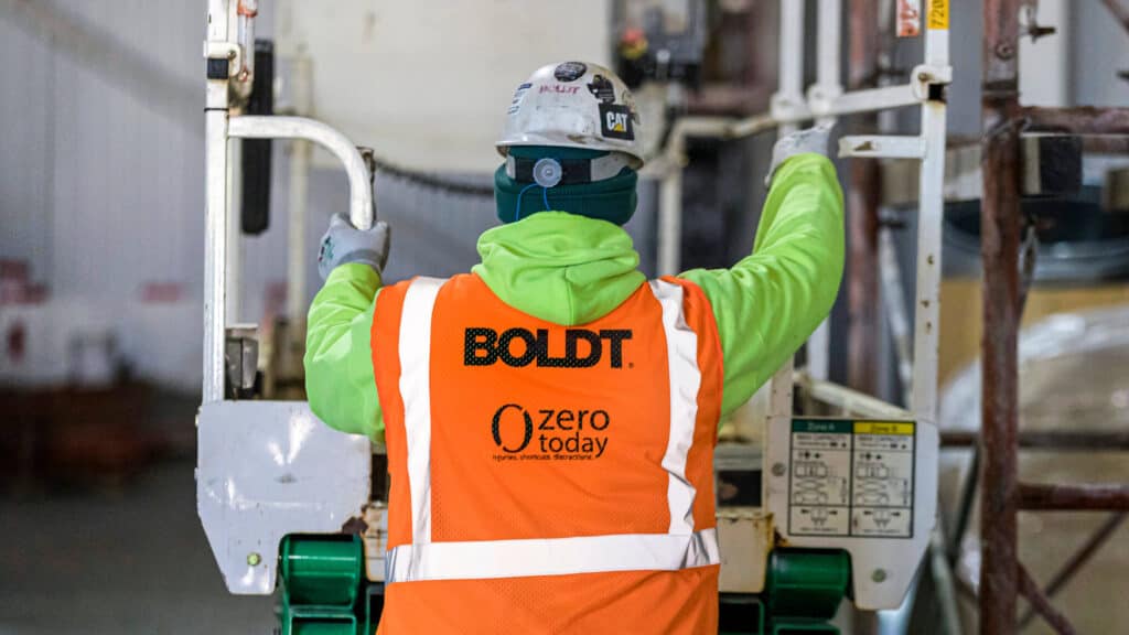  Construction Worker Climbing onto Lift Equipment Wearing Safety Vest with Zero Today Safety Messaging