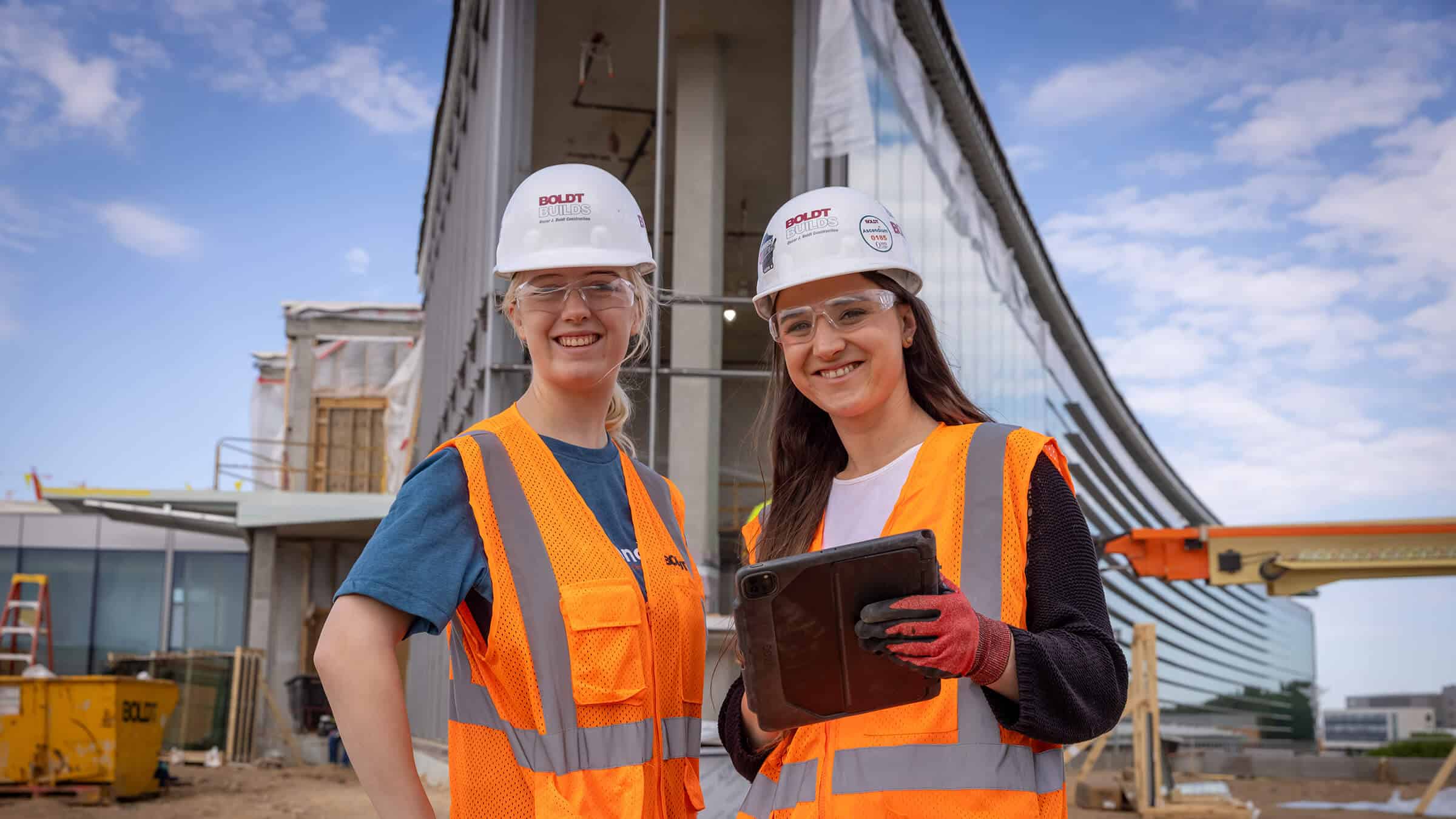 Two young construction women in safety vests and hard hats standing in front of large office building under construction